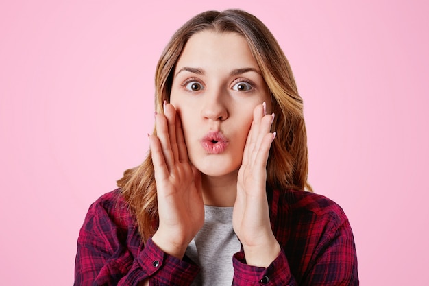 La donna abbastanza adorabile stupita arrotonda le labbra come dice wow, tiene le mani sulle guance, è scioccata, gesti in studio