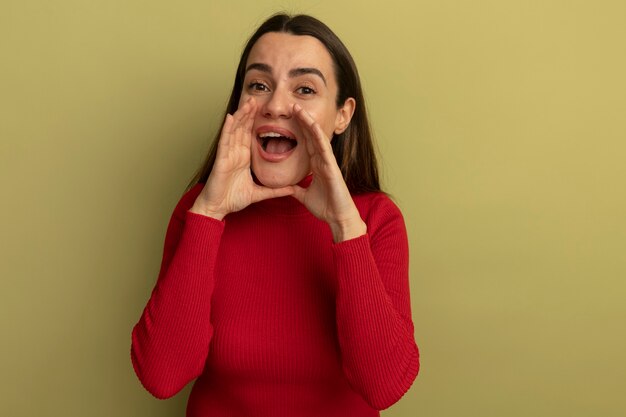 La donna abbastanza caucasica impressionata tiene le mani vicino alla bocca che chiama qualcuno su verde oliva