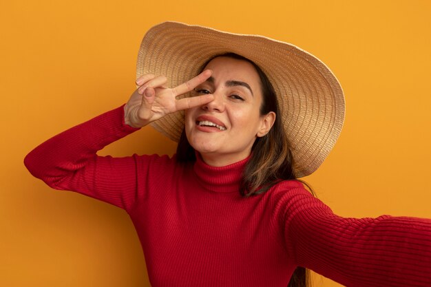 La donna abbastanza caucasica sorridente con il cappello della spiaggia gesticola il segno della mano di vittoria e finge di tenere la macchina fotografica sull'arancio