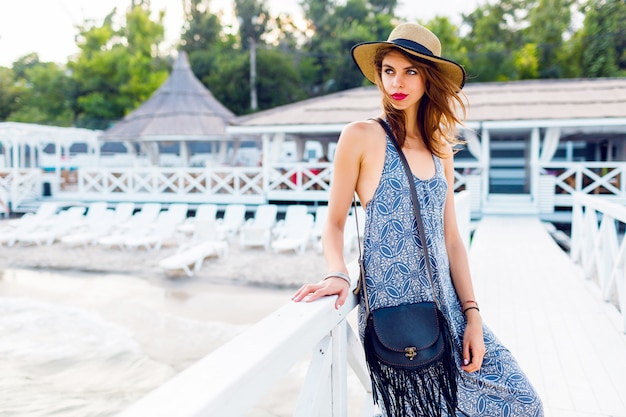 La donna abbastanza esile elegante con il cappello di paglia e la spiaggia lunga si vestono la posa vicino alla località di soggiorno di lusso vicino alla spiaggia. Indossa un'elegante borsa boho.