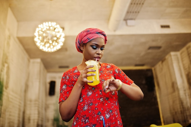 La donna africana alla moda in camicia e cappello rossi ha posato il caffè dell'interno e la limonata bevente dell'ananas