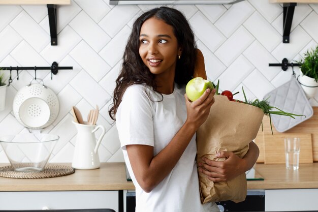 La donna africana che sta sulla cucina tiene un sacco di carta con cibo e mangia una mela