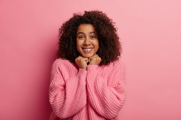 La donna afro dall'aspetto piacevole sorride delicatamente, tiene entrambe le mani sotto il mento, ha una pelle sana, vestita con un maglione invernale, riceve notizie positive, isolata sul muro rosa.