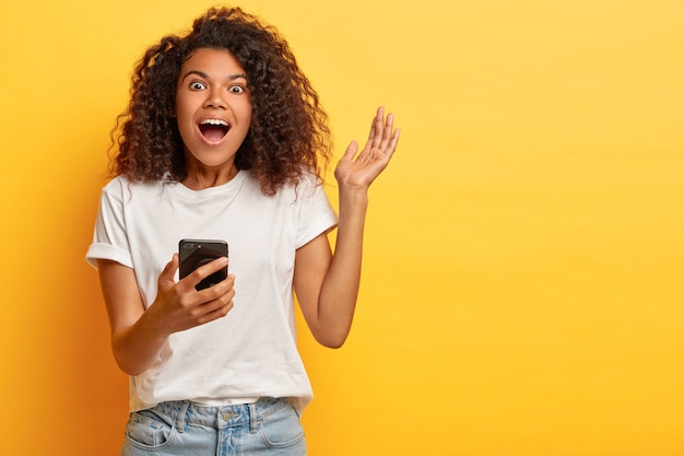 La donna afro emotiva eccitata ride come intrattenuta e divertita, tiene in mano il cellulare moderno, alza il braccio, vestita con una maglietta bianca e jeans, trattiene il respiro dallo stupore, isolato sul muro giallo