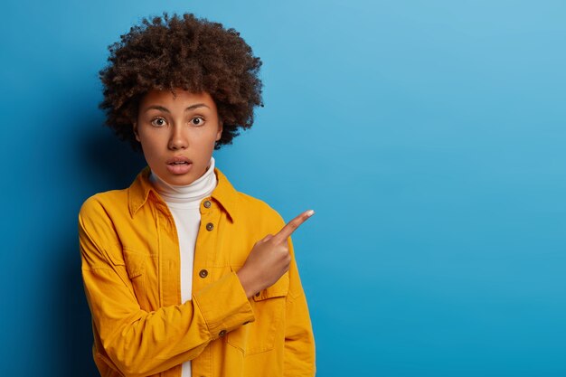 La donna afro scioccata stupita mostra una buona offerta di vendita o un affare, trattiene il respiro, dimostra lo spazio vuoto della copia isolato su sfondo blu.