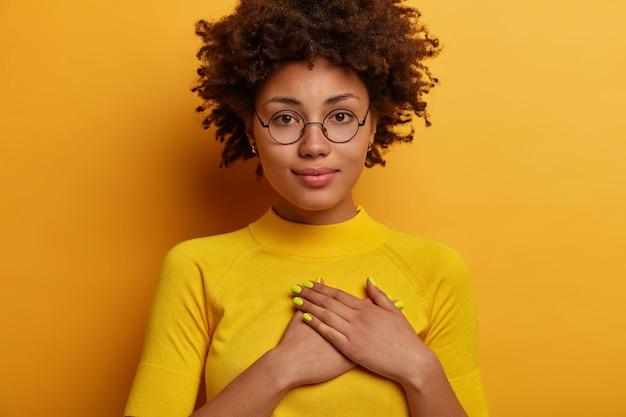 La donna afro seria e calma preme le mani sul cuore in un gesto grato, apprezza le belle parole, esprime gratitudine per il regalo ricevuto, indossa abiti gialli in un colore con il muro, ascolta le congratulazioni