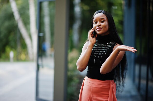 La donna afroamericana alla moda in pantaloni color pesca e camicetta nera parla al telefono all'aperto