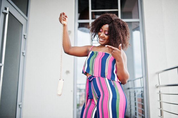 La donna afroamericana alla moda in tuta a righe rosa mostra il dito sulla borsetta e posa