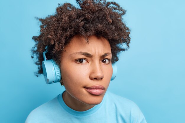 La donna afroamericana dai capelli ricci dispiaciuta seria indossa le cuffie wireless stereo guarda attentamente ascolta musica o audiolibro vestito casualmente isolato sul muro blu.