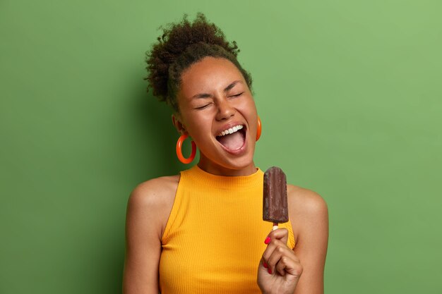 La donna afroamericana felicissima gode del gelato al cioccolato congelato, tiene la bocca aperta, si diverte durante le calde giornate estive, mangia un delizioso dessert, isolato sul muro verde. Cibo spazzatura, calorie