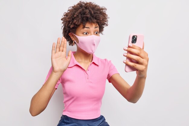 La donna afroamericana in autoisolamento durante la pandemia di coronavirus indossa una maschera protettiva per il viso che fa onde di videochiamata palmo in gesto di saluto mantiene lo smartphone in posizione frontale contro il muro bianco