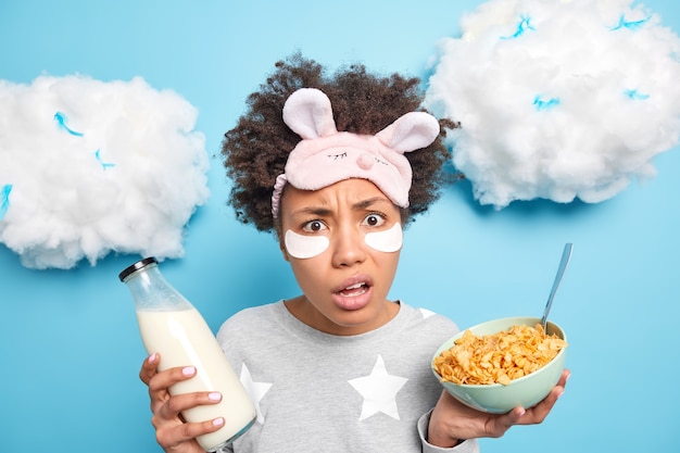 La donna afroamericana perplessa e indignata mangia i cereali per la colazione beve il latte fresco indossa la maschera da notte pigiama vestito isolato sopra il muro blu