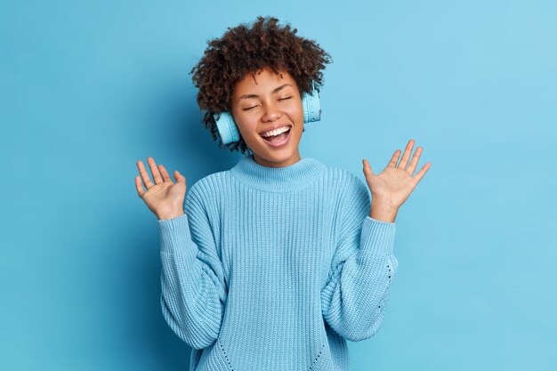 La donna afroamericana positiva con i capelli ricci solleva le palme si diverte mentre ascolta la traccia audio indossa le cuffie wireless vestite con un maglione casual