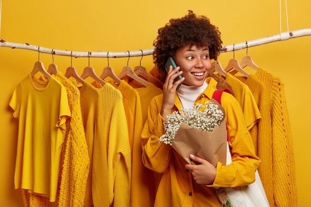 La donna afroamericana positiva si allontana dalla telecamera, ha un'espressione allegra, si oppone alla sbarra dei vestiti, parla al telefono