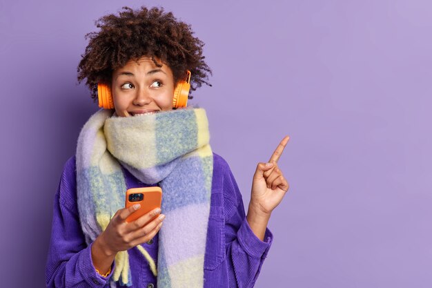 La donna afroamericana premurosa felice avvolta con la sciarpa tiene il telefono cellulare ha comunicazione in linea ascolta la musica tramite i punti delle cuffie stereo nell'angolo in alto a destra mostra lo spazio vuoto per il testo