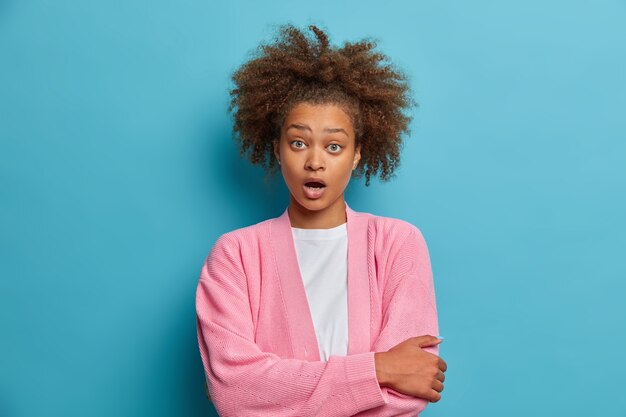 La donna afroamericana scioccata e imbarazzata tiene la bocca ben aperta ha i capelli ricci e folti sente un'incredibile novità fissa stupita vestita con un maglione rosa casual.