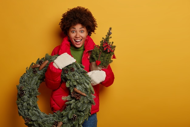 La donna afroamericana stupita sorpresa tiene la corona verde e l'abete, ha un'espressione gioiosa, si prepara per Natale o Capodanno, isolato sopra il muro giallo.