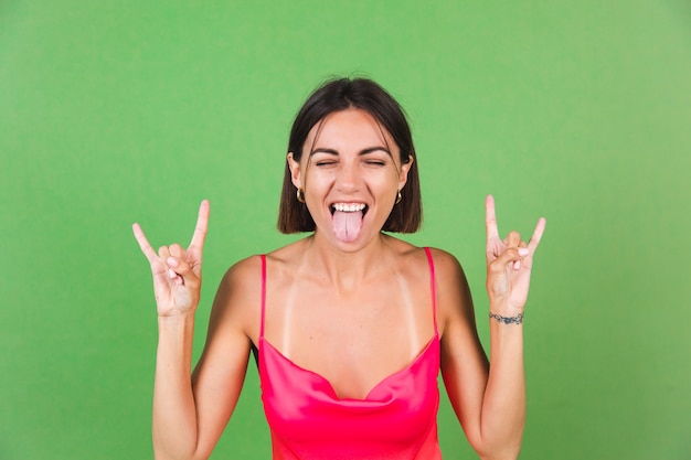 La donna alla moda in vestito di seta rosa isolata su verde con il gesto della roccia mostra la sua lingua, gioiosa divertente