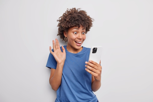 La donna allegra con i capelli ricci fa selfie o fa le onde di videochiamata ciao il gesto ha sorpreso l'espressione felice sente grandi notizie indossa una maglietta blu casual isolata su sfondo bianco