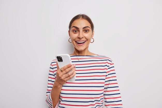 La donna allegra con un aspetto piacevole tiene il telefono cellulare gode della comunicazione online indossa un maglione a righe casual isolato su un muro bianco