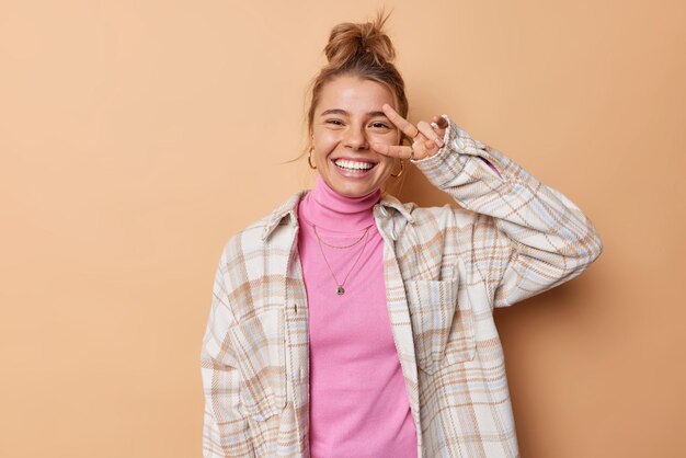 La donna allegra e contenta mostra il gesto di vittoria o di pace sugli occhi sorride con gioia indossa un dolcevita rosa e una camicia a scacchi isolata su sfondo beige. Concetto di linguaggio del corpo ed emozioni positive