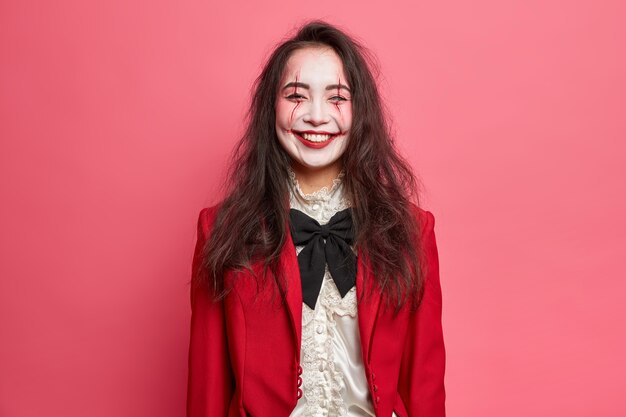 La donna allegra e spaventosa con il trucco di Halloween ha la faccia pallida indossa il costume per la festa di carnevale in posa contro il muro rosa in vacanza