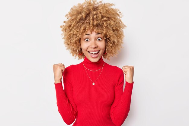 La donna allegra emotiva con i capelli ricci folti trionfa su qualcosa che stringe i pugni fissa i sorrisi della fotocamera indossa un dolcevita rosso casual isolato su sfondo bianco. Evviva l'ho fatto