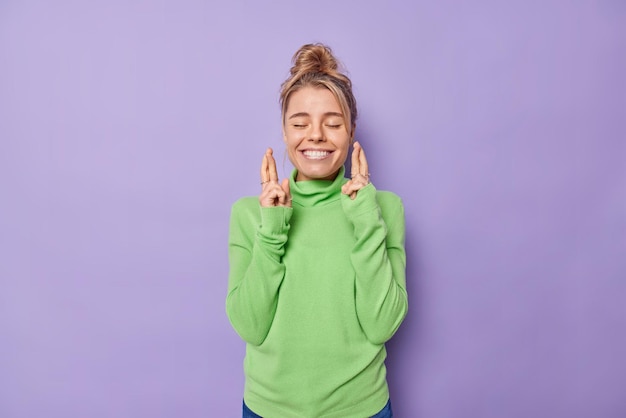 La donna allegra sta con gli occhi chiusi sorride boadly fa il desiderio incrocia le dita crede nella buona fortuna o la fortuna indossa un maglione verde casual isolato su sfondo viola. Possano i miei sogni diventare realtà