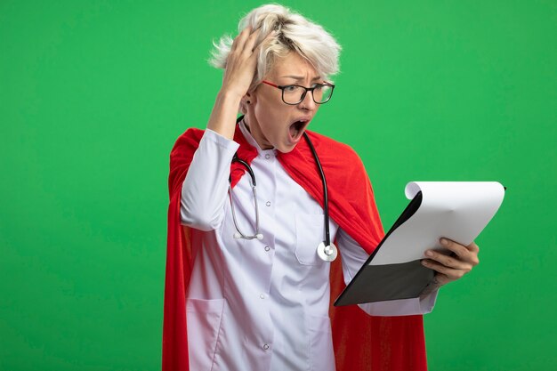 La donna ansiosa del supereroe slavo in uniforme del medico con il mantello rosso e lo stetoscopio in vetri ottici mette la mano sulla testa e guarda gli appunti isolati sulla parete verde con lo spazio della copia