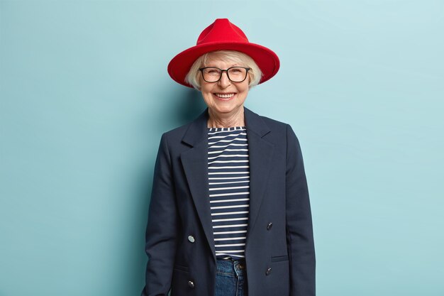 La donna anziana alla moda alla moda sorride felicemente, mostra i denti bianchi, ha la pelle rugosa, vestita con abiti formali eleganti, è di buon umore, pronta per il lavoro, gode di una bella giornata, isolata sul muro blu