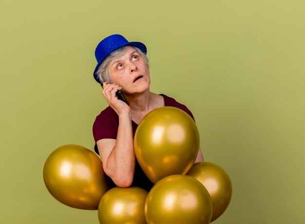 La donna anziana scioccata che porta il cappello del partito tiene i palloni dell'elio che parlano sul telefono che osserva in su isolato sulla parete verde oliva