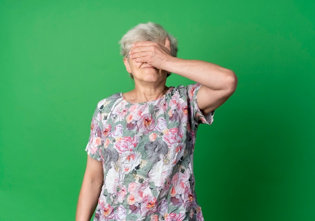 La donna anziana scontenta chiude gli occhi con la mano isolata sulla parete verde