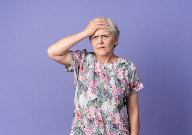 La donna anziana scontenta mette la mano sulla fronte che guarda in avanti isolata sulla parete viola