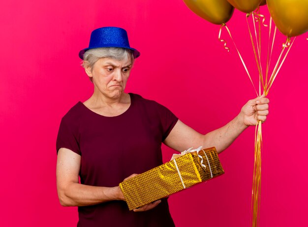 La donna anziana senza tracce che porta il cappello del partito tiene i palloni dell'elio e guarda la confezione regalo sul rosa