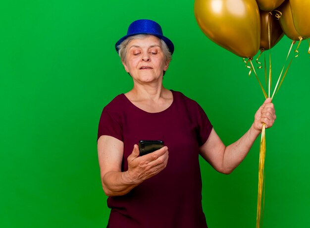 La donna anziana sicura che porta il cappello del partito tiene i palloni dell'elio e guarda il telefono sul verde