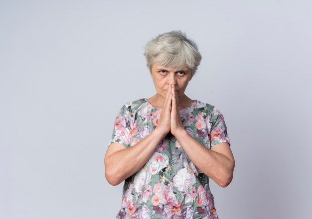 La donna anziana sicura tiene le mani insieme vicino alla bocca isolata sulla parete bianca
