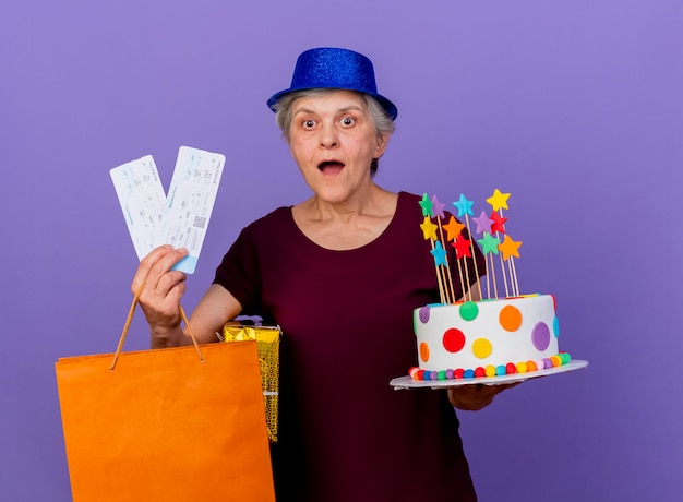 La donna anziana sorpresa che indossa il cappello del partito tiene il sacchetto della spesa della carta della scatola regalo dei biglietti aerei e la torta di compleanno isolata sulla parete viola con lo spazio della copia