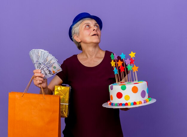 La donna anziana sorpresa che indossa il cappello del partito tiene il sacchetto della spesa di carta del contenitore di regalo dei soldi e la torta di compleanno che esamina il lato isolato sulla parete viola con lo spazio della copia