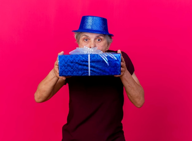 La donna anziana sorpresa che porta il cappello del partito tiene ed esamina la macchina fotografica sopra il contenitore di regalo sul colore rosa