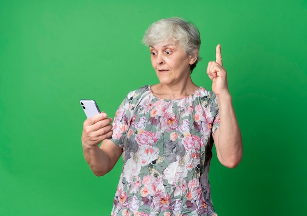 La donna anziana sorpresa esamina il telefono rivolto verso l'alto isolato sulla parete verde