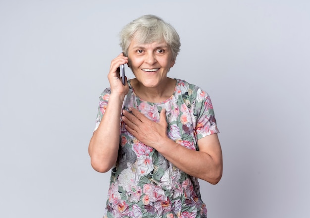 La donna anziana sorridente mette la mano sul mento che parla sul telefono isolato sulla parete bianca