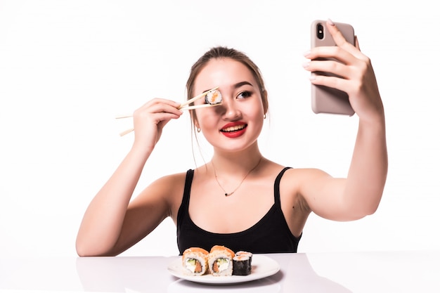 La donna asiatica abbastanza sorridente copre il suo occhio di rotolo di sushi e fa il selfie sul suo telefono