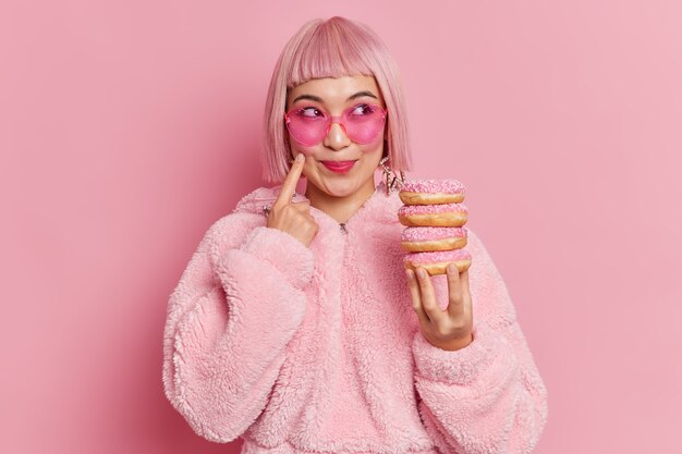 La donna asiatica abbastanza sorridente sweettooth con capelli rosa alla moda indossa occhiali da sole e pelliccia detiene un mucchio di ciambelle pone sogni al coperto su qualcosa. Colpo monocromatico. La femmina ama mangiare le ciambelle