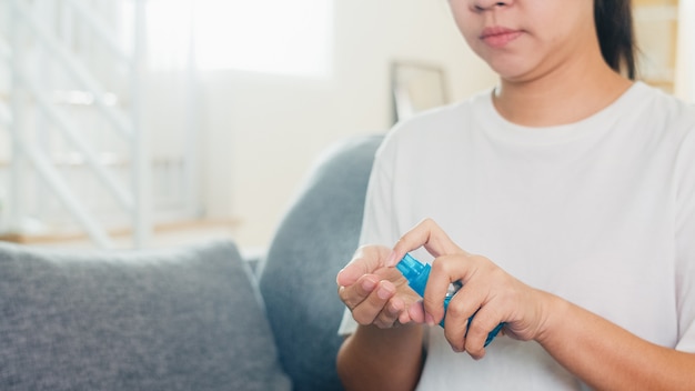 La donna asiatica che usando la mano del lavaggio del disinfettante della mano dello spruzzo dell'alcool per protegge il coronavirus. Femmina che si siede alla mano pulita del divano per l'igiene quando il distanziamento sociale resta a casa e il tempo di auto-quarantena.