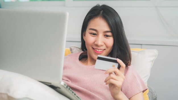 La donna asiatica che utilizza il commercio elettronico di acquisto della carta di credito e del computer portatile, femmina si rilassa sentendosi l'acquisto online felice che si siede sul sofà nel salone a casa. Le donne di stile di vita si rilassano a casa concetto.