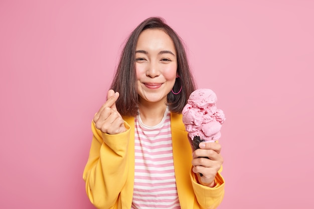 La donna asiatica compiaciuta fa un mini gesto del cuore come un segno coreano che tiene un delizioso gelato mangia un dolce gustoso dessert congelato indossa una maglietta a righe e una giacca gialla posa contro il muro rosa. Estate