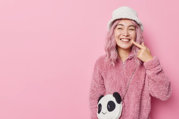 La donna asiatica dai capelli piuttosto rosa indica i punti del sorriso a trentadue denti sui denti bianchi indossa capispalla alla moda che guarda volentieri lontano isolato su sfondo rosa spazio vuoto per la tua promozione