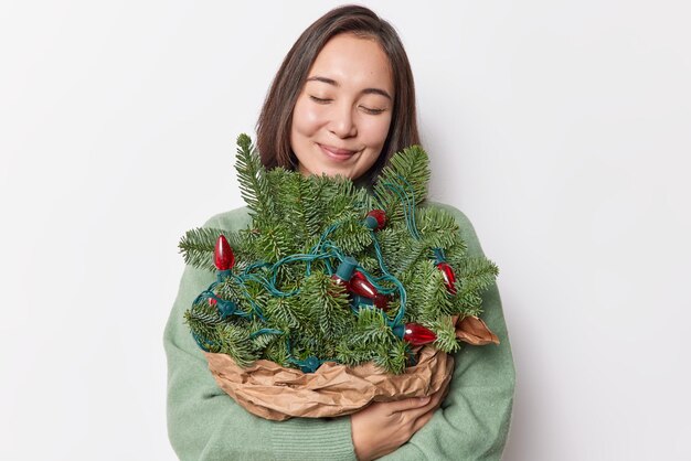 La donna asiatica dai capelli scuri soddisfatta abbraccia il bouquet di rami di abete decorati con ghirlanda retrò indossa un maglione casual tiene gli occhi chiusi sogna ad occhi aperti su qualcosa di isolato su sfondo bianco