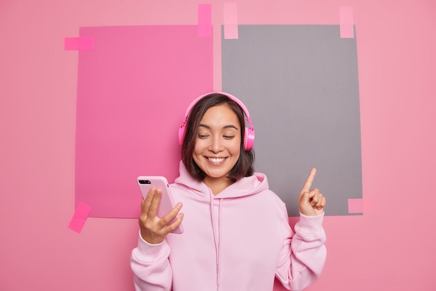 La donna asiatica millenaria soddisfatta fa una videochiamata che promuove qualcosa che indica in uno spazio vuoto sorride piacevolmente mostra la direzione di vendita logo banner del negozio indossa pose con cappuccio contro il muro rosa