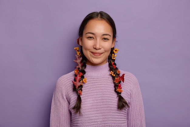 La donna asiatica naturale felice celebra l'autunno, ha due trecce decorate con foglie autunnali rosse, bacche e fiori, indossa un maglione viola, isolato sul muro viola. Tempo di ottobre